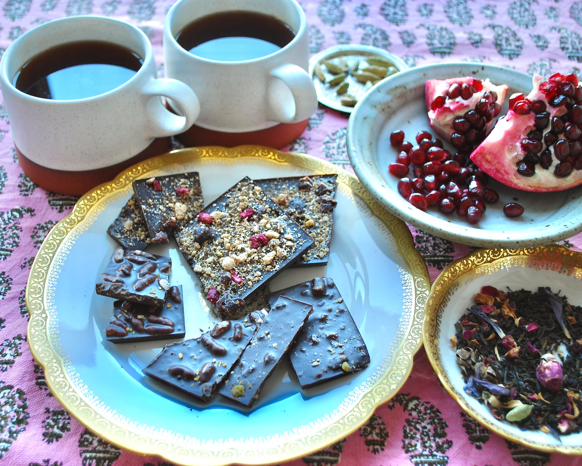 A set of gourmet dark chocolate bars arranged on a plate with cups of coffee and bowls of pomegranate seeds and tea leaves in the background, all presented on a table with a pink tablecloth.
