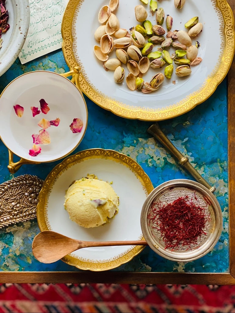 Saffron Ice Cream Scoop in a porcelain bow surrounded by plates of rose petals, pistachios and saffron with a wooden spoon