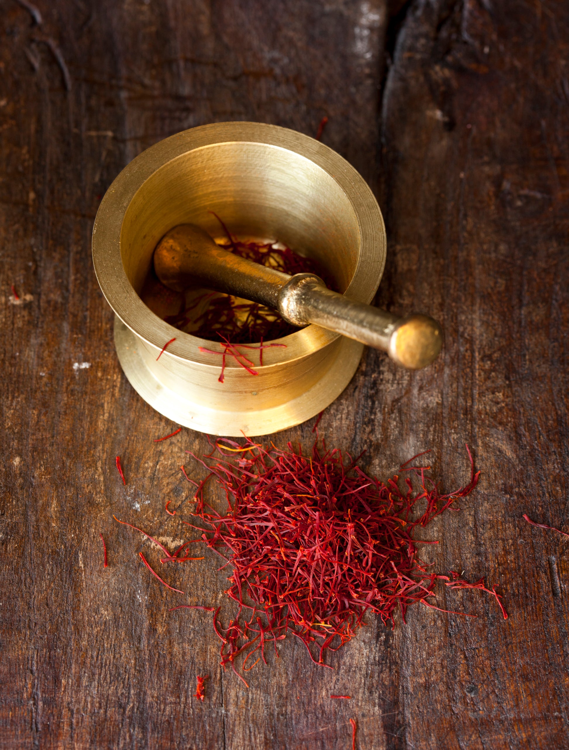 A brass mortar and pestle with a pile of red saffron threads on a wooden surface.