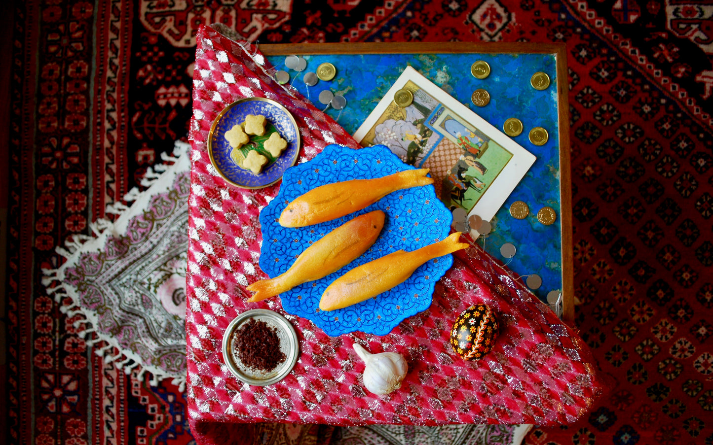 Nowruz setup featuring marzipan goldfish, gold coins, garlic, spices and cookies on a tapestry