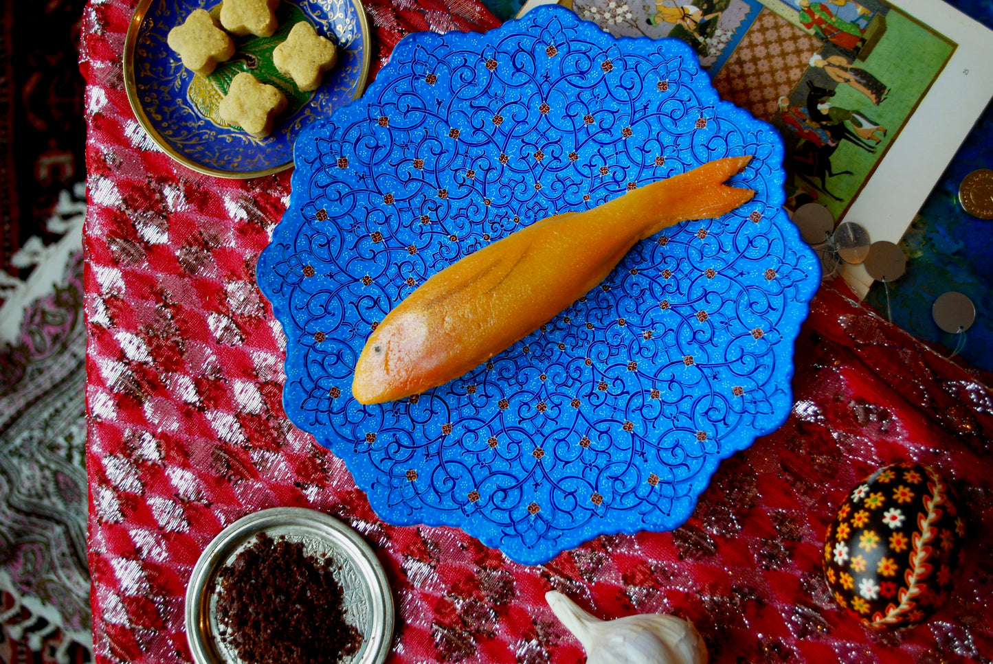 A single marzipan goldfish on a blue plate surrounded by chickpea cookies and spices